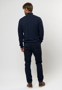 Marineblauer Strickpullover mit hohem Kragen, kombiniert mit dunkelblauen Jeans und braunen Lederschuhen, von hinten betrachtet.