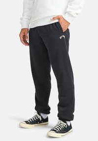 Zwarte sweatpants met elastische boorden, voorzien van een klein gedrukt logo. Gecombineerd met zwart-witte sneakers en een lichtgrijs sweatshirt.
