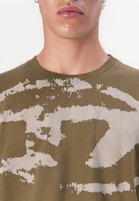 Olivově zelené bavlněné tričko s poškozeným světle béžovým vzorem, kulatým výstřihem a krátkými rukávy, zdůrazňující texturovanou tkaninu.