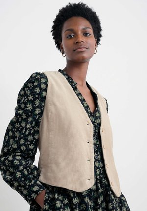 Beige corduroy vest met vijf knopen over een donker bloemenjurk met pofmouwen. Gelaagde stof met verticale ribbelpatronen.