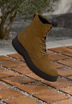 Botte montante à lacets en daim couleur tan avec semelle noire, en lévitation au-dessus d'un bloc de bois et d'un sol à motif de galets en extérieur.