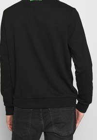 Felpa nera con collo rotondo e maniche lunghe; presenta orlo e polsini a righe. Sul retro è visibile un testo del logo verde vicino al colletto.