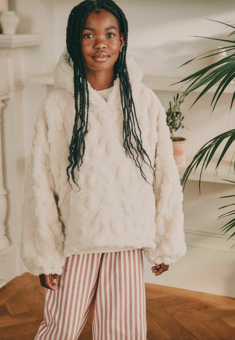 Haut à capuche en polaire couleur crème avec un motif texturé, poche avant et un large col en V. Associé à un pantalon évasé rayé rouge et blanc.