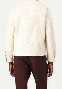 Broderad krämfärgad jacka med blommönster och textilstruktur, med krage och knäppärmar, bärs över bruna byxor.