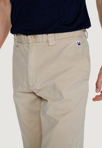 Pantaloni in cotone beige con una texture liscia, dotati di chiusura con bottone e zip e di una piccola toppa logo in rosso, bianco e blu sulla cintura.