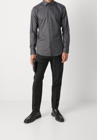 Grijs overhemd met lange mouwen en een button-down kraag, gecombineerd met zwarte op maat gemaakte broek en zwarte leren schoenen, met een zwarte portemonnee in de hand.