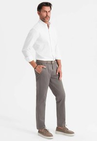 Camicia bianca a maniche lunghe con colletto a camicia, pantaloni grigi, sneaker grigie chiare con lacci, mani in tasca, sfondo uniforme.