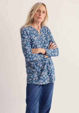 Kobieta z długimi blond włosami, ubrana w niebieską, kwiecistą bluzkę z długim rękawem i niebieskie dżinsy, stoi z założonymi rękami na tle jednolitego tła.