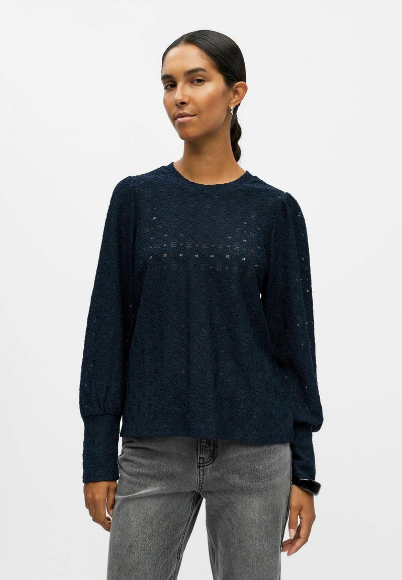 Haut en dentelle bleu marine à manches longues avec un motif texturé, un col rond et des manches froncées, assorti à un jean gris clair.