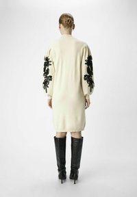 Robe en tricot écrue avec broderie florale noire sur les manches. Présente des manches bouffantes surdimensionnées et est associée à des bottes noires montantes.