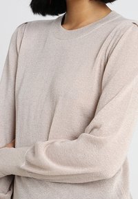 Ljusrosa tröja i ett mjukt, texturerat material med rundad halsringning och lätt puffade ärmar. Inga synliga mönster eller detaljer i hårdvara.