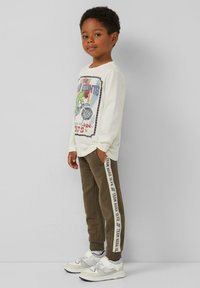 Weißes Langarmshirt mit buntem Grafikdruck, olivgrüne Sweatpants mit seitlichem Streifen und helle Sneaker mit Mesh-Elementen.