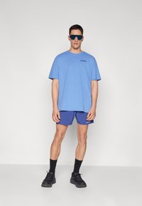 Tricou din bumbac albastru cu logo negru, asortat cu pantaloni scurți purpurii. Modelul poartă ochelari de soare reflectanți albaștri și încălțăminte sport neagră.