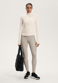 Lightweight, beige puffer vest with vertical quilted sections, paired with beige leggings featuring teal side stripes, black sneakers, and a black tote bag.