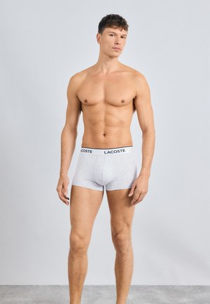 Gray cotton boxer shorts with a black Lacoste logo on the waistband, fitted design, and smooth texture, modeled by a standing individual.