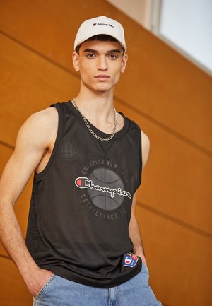 Camiseta de baloncesto de malla negra con gráfico de logo, combinada con pantalones cortos de mezclilla azul claro. El modelo lleva una gorra blanca de Champion y un collar de cadena plateada.
