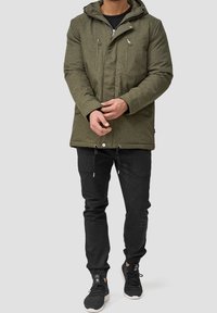 Olivgrüne Parkajacke aus strukturiertem Material, mit durchgehendem Reißverschluss, zwei Brusttaschen und einer verstellbaren Kapuze. Kombiniert mit schwarzen Hosen und Sneakers.