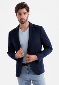 Marineblauer Blazer mit Reverskragen, Ein-Knopf-Verschluss und zwei Fronttaschen, über einem hellblauen V-Ausschnitt-Pullover und hellblauen Jeans getragen.