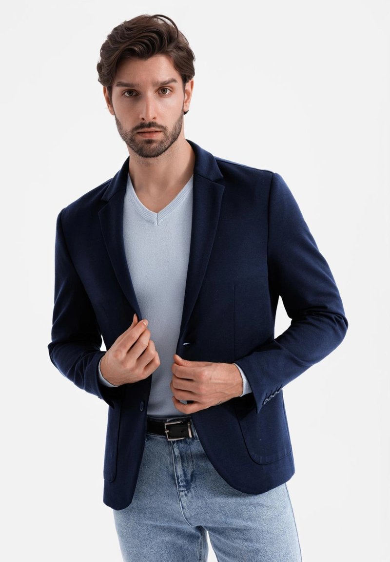 Marineblauer Blazer mit Reverskragen, Ein-Knopf-Verschluss und zwei Fronttaschen, über einem hellblauen V-Ausschnitt-Pullover und hellblauen Jeans getragen.