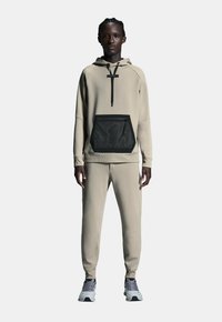 Lichtbeige hoodie met een zwart gaaszak, gecombineerd met bijpassende beige broek. Voorzien van een ritssluiting aan de voorkant en ribgebreide manchetten. Geweven stof.