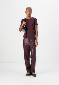 Chemise maroon à manches courtes, associée à un pantalon en cuir maroon brillant, un sac à main noir et des ballerines à imprimé léopard, sur un fond blanc uni.