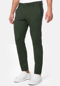 Dark green, slim-fit pants made from a smooth fabric with a flat front, side pockets, and a waistband with a button closure, paired with white sneakers.