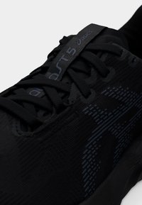 Τα μαύρα αθλητικά παπούτσια διαθέτουν πλεκτό επάνω μέρος με υφή, μαύρα κορδόνια και διακριτικό μπλε σχέδιο στο πλάι. Το λογότυπο είναι ορατό στη γλώσσα του παπουτσιού.