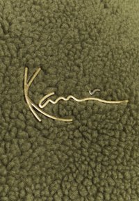 Oliven grønn fleece-stoff med en teksturert overflate, med en metallisk gulllogo som ligner flytende linjer og en gren-design.
