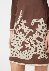 Vestido marrón con intrincados bordados en crema que presentan formas y patrones abstractos, finalizando con un dobladillo con flecos.