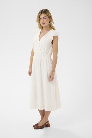 Nő fehér, ujjatlan, midi hosszúságú, kivágásos részletekkel díszített ruhában és barna, lapos sarkú fonott cipőben, egyszerű háttér előtt áll.