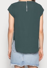 Femme portant un haut à manches courtes vert foncé avec une fermeture éclair au dos et une jupe en denim bleu clair effilochée, vue de derrière.