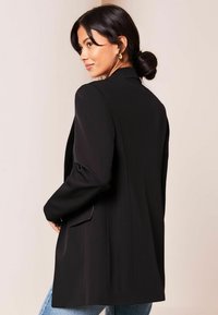 Blazer negro con hombros ajustados, mangas ligeramente acolchadas y una abertura en la parte posterior. Tejido suave y diseño a medida.