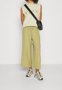 Person med ärmlös beige toppen med gul grafisk design, lösa ljusgröna byxor, beige sneakers och svart axelväska på vit bakgrund.