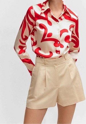 Femme portant un short plissé beige taille haute et une chemise blanche à boutons avec des motifs abstraits rouges audacieux, debout les mains derrière le dos.