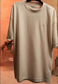 Bež oversize majica kratkih rukava s malim izvezenim logotipom, koja visi na crnoj vješalici na narančastoj pozadini s biljkama i namještajem.