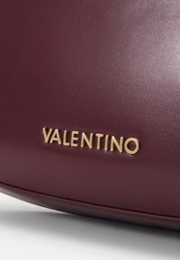 Burgundy läderhandväska med en slät yta, som har en framträdande guld "VALENTINO"-logotyp. Designen är elegant och strömlinjeformad.