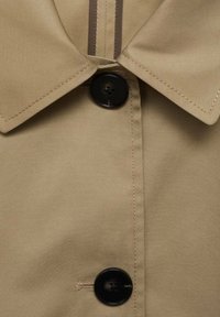 Beige stoffen jas met een klassieke kraag, zwarte knopen en contrasterende stiksels langs de sluiting, met een gladde textuur.