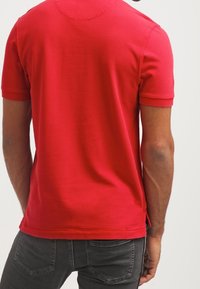 Homme portant un polo rouge uni à manches courtes et un jean gris foncé, vu de dos sur un fond uni.