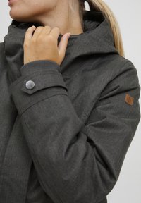 Donkergrijze jas met een hoge kraag, gestructureerde stof en een leren patch. Voorzien van een metalen drukknoopsluiting op de mouw voor een aanpasbare pasvorm.