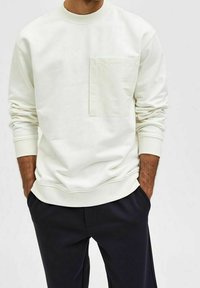 Vit crewneck sweatshirt i mjukt tyg, med en frontficka och ribbad mudd, i kombination med mörka byxor.