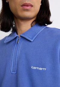 Blue polo shirt with a zip collar, textured fabric, and white Carhartt logo on the left side. Features a relaxed fit and clean lines.