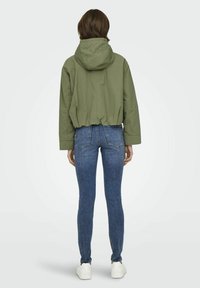 Veste olive verte coupée avec capuche, associée à un jean bleu ajusté. La veste a une texture légère et lisse. Des baskets blanches complètent la tenue.
