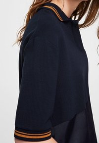 Haut en maille bleu marine avec un col bandeau, présentant des rayures orange contrastantes sur le col et les manches ; coupe décontractée et tissu texturé.