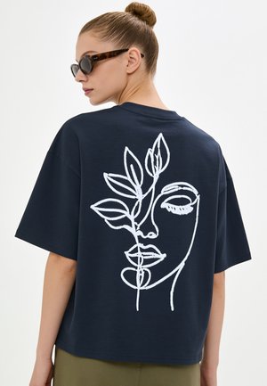 Femme portant un t-shirt oversize bleu foncé avec un dessin au trait blanc d’un visage et d’une plante dans le dos, des lunettes de soleil et les cheveux attachés en chignon.