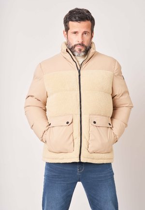 Homme avec barbe portant une polaire sherpa beige et une veste matelassée avec poches avant, debout les mains dans les poches devant un fond uni.