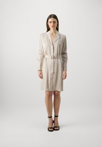 Beige selyem ingruha hosszú ujjal, hajtott gallérral, elöl gombokkal és derékban illeszkedő övvel. Fekete magassarkú cipővel egészül ki a megjelenés.