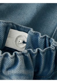 Elastīga zila džinsu auduma monta ar caurspīdīgu pogu, kas pievienota baltai regulējamai siksnai iekšā apģērba jostasvietā.