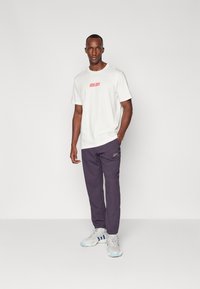 Tricou alb cu text roșu, combinat cu pantaloni de jogging violeți. Modelul poartă adidași gri și albastri. Design simplu, potrivire lejeră.