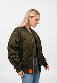 Giubbotto bomber verde oliva con texture trapuntata, polsini a coste e chiusura con zip; presenta un'etichetta arancione a contrasto sull'orlo.