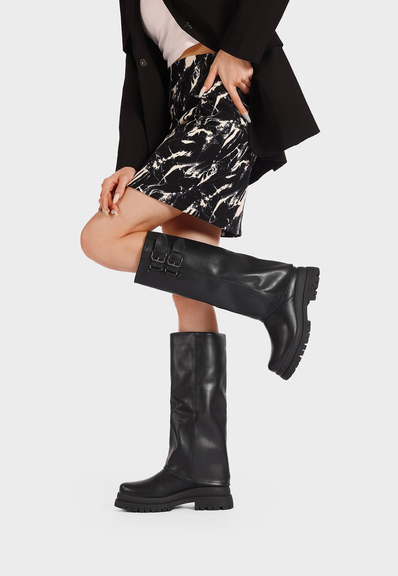 Botas de cuero negro hasta la rodilla con una suela gruesa, adornadas con dos hebillas en el lateral y una textura lisa, usadas con una falda estampada.